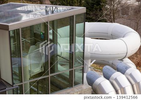 Sleek architectural glass building with a thrilling white curving water slide, inviting visitors to enjoy modern aquatic leisure, summer fun, and park entertainment Sleek architectural glass building with a thrilling white curving water slide, inviting visitors to enjoy modern aquatic leisure, summer fun, and park entertainment 139412566
