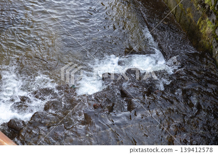 Dynamic stream water vigorously flowing over dark river rocks, generating frothy white bubbles, representing nature's refreshing purity and ecological vitality for environmental concepts Dynamic stream water vigorously flowing over dark river rocks, generating frothy white bubbles, representing nature's refreshing purity and ecological vitality for environmental concepts 139412578