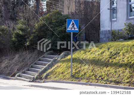 Blue pedestrian crossing sign marks safe public pathway by rustic stone stairs on a sunny green slope, promoting urban pedestrian safety Blue pedestrian crossing sign marks safe public pathway by rustic stone stairs on a sunny green slope, promoting urban pedestrian safety 139412588