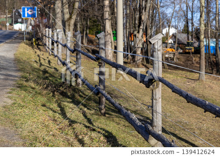 Rustic wooden fence with rough logs and wire defines a sunny country road boundary, leading towards distant village houses and bare forest trees Rustic wooden fence with rough logs and wire defines a sunny country road boundary, leading towards distant village houses and bare forest trees 139412624