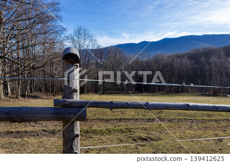 Rustic wooden fence with electric wire marking a winter field boundary, showcasing bare trees and distant mountains under a bright blue sky, portraying rural nature Rustic wooden fence with electric wire marking a winter field boundary, showcasing bare trees and distant mountains under a bright blue sky, portraying rural nature 139412625
