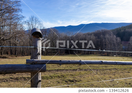 Weathered wooden fence with electric wires delineating a calm rural field, featuring bare winter trees and distant mountains under a blue sky Weathered wooden fence with electric wires delineating a calm rural field, featuring bare winter trees and distant mountains under a blue sky 139412626