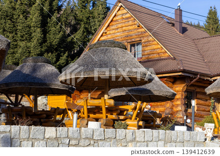 Traditional wooden log cabin lodge with rustic outdoor seating and thatched straw umbrellas, creating an inviting atmosphere for a serene mountain resort vacation Traditional wooden log cabin lodge with rustic outdoor seating and thatched straw umbrellas, creating an inviting atmosphere for a serene mountain resort vacation 139412639