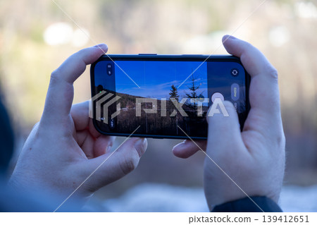 Hands holding a smartphone, actively capturing an outdoor landscape, displaying the camera app with PHOTO mode, symbolizing travel, hobby, and digital content creation Hands holding a smartphone, actively capturing an outdoor landscape, displaying the camera app with PHOTO mode, symbolizing travel, hobby, and digital content creation 139412651