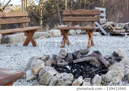 Rustic stone fire pit with ash and wood on a gravel patio, featuring wooden benches, perfect for outdoor social gathering and relaxation Rustic stone fire pit with ash and wood on a gravel patio, featuring wooden benches, perfect for outdoor social gathering and relaxation 139412660