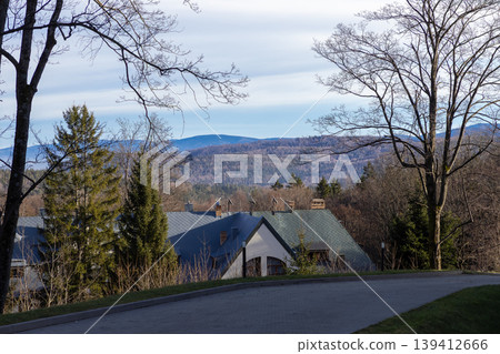 Panoramic mountain landscape featuring vast forest and distant residential rooftops, framed by bare trees under a bright blue sky, symbolizing peaceful nature and serene rural retreat Panoramic mountain landscape featuring vast forest and distant residential rooftops, framed by bare trees under a bright blue sky, symbolizing peaceful nature and serene rural retreat 139412666