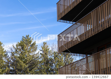 Modern residential building with contemporary balconies and brown vertical railings, framed by green trees against a clear blue sky, ideal for urban architecture Modern residential building with contemporary balconies and brown vertical railings, framed by green trees against a clear blue sky, ideal for urban architecture 139412667