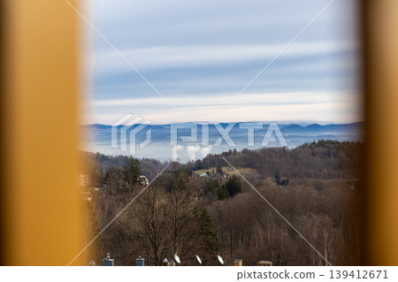 Framed scenic view reveals a foggy winter valley with distant industrial smoke emerging above bare tree-covered hills and layered blue mountains Framed scenic view reveals a foggy winter valley with distant industrial smoke emerging above bare tree-covered hills and layered blue mountains 139412671