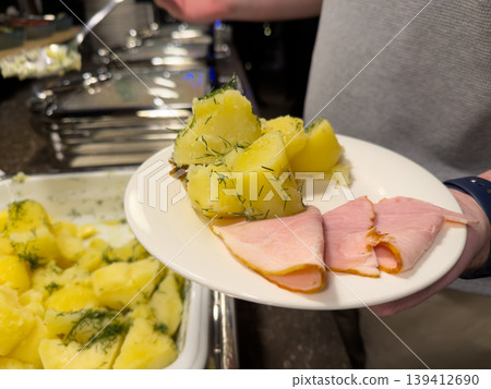 Person holds a white plate featuring savory boiled potatoes, seasoned with fresh dill, alongside delicate slices of ham for a casual buffet meal Person holds a white plate featuring savory boiled potatoes, seasoned with fresh dill, alongside delicate slices of ham for a casual buffet meal 139412690