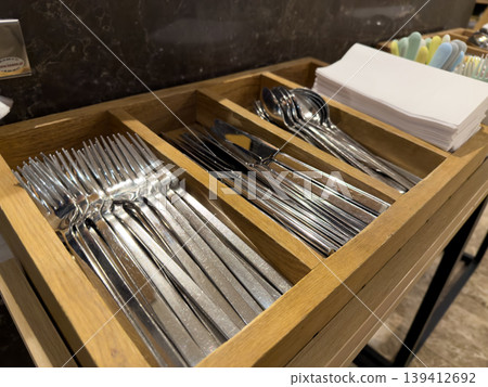 Clean, polished stainless steel forks, knives, and spoons are perfectly organized within a sleek wooden holder, with white napkins, for buffet service and elegant dining experiences Clean, polished stainless steel forks, knives, and spoons are perfectly organized within a sleek wooden holder, with white napkins, for buffet service and elegant dining experiences 139412692