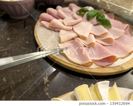 Serving tongs pick a slice of fresh deli ham from a vibrant platter with assorted cold cuts and basil, perfect for a buffet or breakfast spread 139412694