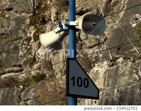 Two robust public address loudspeakers mounted on a blue pole featuring a 100 sign, for critical announcements in a rugged natural environment Two robust public address loudspeakers mounted on a blue pole featuring a 100 sign, for critical announcements in a rugged natural environment 139412701