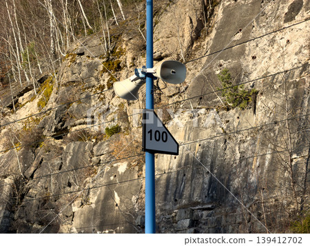 White loudspeakers and a 100 sign on a blue pole, indicating a public address system in a rugged rocky cliff environment for safety warnings White loudspeakers and a 100 sign on a blue pole, indicating a public address system in a rugged rocky cliff environment for safety warnings 139412702
