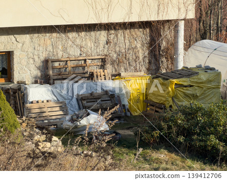 Stacks of construction materials and wooden pallets covered with tarps stored next to a building wall, indicating a renovation or home improvement project Stacks of construction materials and wooden pallets covered with tarps stored next to a building wall, indicating a renovation or home improvement project 139412706