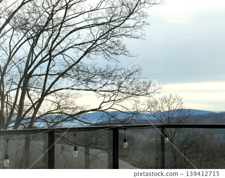 Bare tree branches frame a serene mountain landscape and overcast sky, viewed from a modern balcony with string lights, evoking peace and natural beauty 139412715