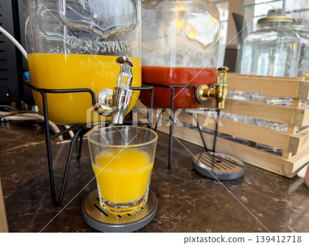 Glass beverage dispenser pouring fresh orange juice into a glass, alongside another dispenser, offering refreshing drink options at a breakfast buffet 139412718