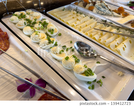 Platter of creamy deviled eggs topped with fresh green onion garnish, offered alongside diverse gourmet cheeses at a vibrant breakfast buffet 139412723