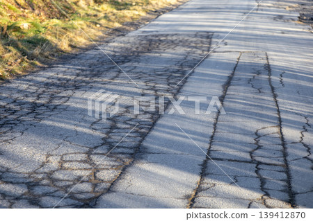 Deteriorated asphalt road displaying deep cracks and wear, bathed in sunlight and contrasting shadows, symbolizing infrastructure neglect and urgent repair Deteriorated asphalt road displaying deep cracks and wear, bathed in sunlight and contrasting shadows, symbolizing infrastructure neglect and urgent repair 139412870