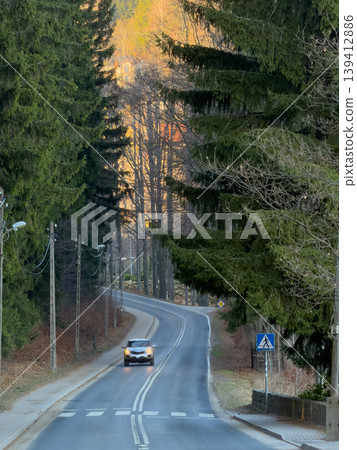 White car drives on a winding asphalt road through a dense forest, featuring a pedestrian crossing sign amidst tall pine trees, highlighting nature's journey White car drives on a winding asphalt road through a dense forest, featuring a pedestrian crossing sign amidst tall pine trees, highlighting nature's journey 139412886