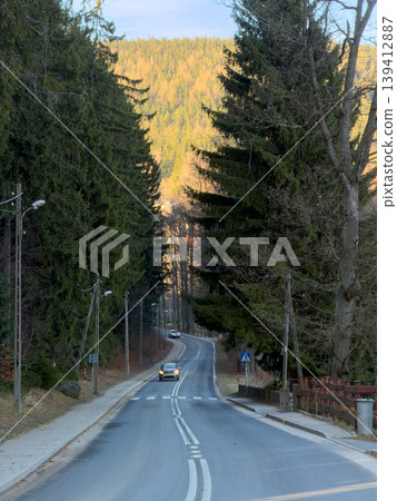 Winding road with cars driving through a scenic mountain forest, flanked by tall evergreen trees, showing a tranquil journey with distant golden foliage Winding road with cars driving through a scenic mountain forest, flanked by tall evergreen trees, showing a tranquil journey with distant golden foliage 139412887