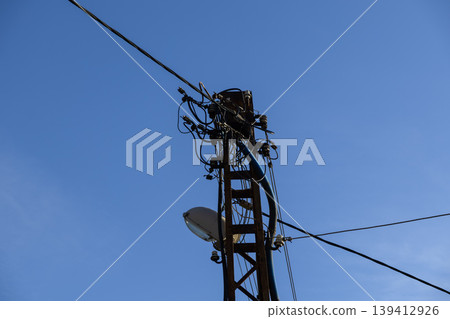 Rusted electricity pole featuring a complex web of tangled power lines and an urban street light, conveying essential energy infrastructure against a clear blue sky Rusted electricity pole featuring a complex web of tangled power lines and an urban street light, conveying essential energy infrastructure against a clear blue sky 139412926