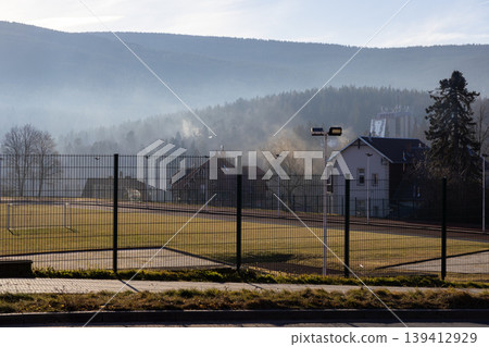 Rural fenced green sports field with soccer goalposts, showcasing distant misty mountain forests and village houses under hazy morning sky, conveying peaceful atmosphere Rural fenced green sports field with soccer goalposts, showcasing distant misty mountain forests and village houses under hazy morning sky, conveying peaceful atmosphere 139412929