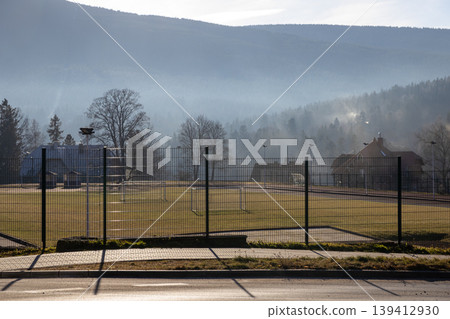 A serene landscape view of an empty soccer field with white goals behind a fence, featuring a hazy mountain forest and a peaceful outdoor setting A serene landscape view of an empty soccer field with white goals behind a fence, featuring a hazy mountain forest and a peaceful outdoor setting 139412930
