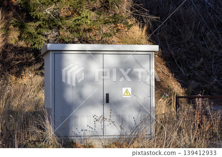 Gray outdoor electrical cabinet, displaying a lightning bolt warning sign, stands amidst dry grass, symbolizing crucial utility and power distribution infrastructure 139412933
