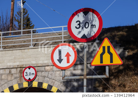 Multiple road signs display a 3,0 m height restriction under a bridge, alongside warnings for narrow passages and traffic priority, promoting road safety Multiple road signs display a 3,0 m height restriction under a bridge, alongside warnings for narrow passages and traffic priority, promoting road safety 139412964