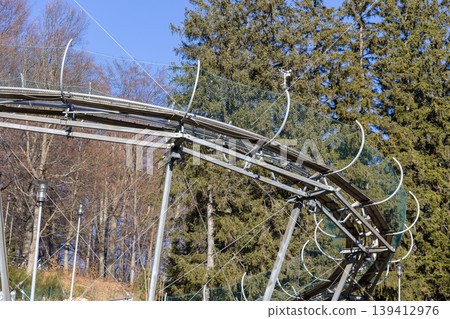 Elevated metal alpine coaster track with robust safety nets curving through a serene mountain forest, symbolizing thrilling outdoor recreation adventure Elevated metal alpine coaster track with robust safety nets curving through a serene mountain forest, symbolizing thrilling outdoor recreation adventure 139412976