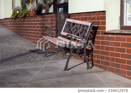 Vintage wrought iron bench with light pink wooden slats casts a sharp shadow on the sidewalk by a textured brick wall, offering public relaxation and urban charm Vintage wrought iron bench with light pink wooden slats casts a sharp shadow on the sidewalk by a textured brick wall, offering public relaxation and urban charm 139412978