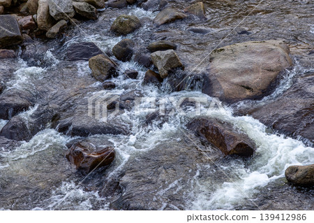 Clear river water flows vigorously among dark wet rocks, creating foamy white rapids and swirling patterns, representing nature's dynamic energy and persistent force Clear river water flows vigorously among dark wet rocks, creating foamy white rapids and swirling patterns, representing nature's dynamic energy and persistent force 139412986