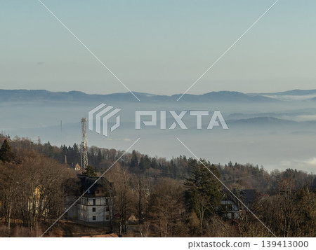 Mountain landscape with a communication tower rising above houses and a valley filled with dense fog, creating a serene and expansive morning view Mountain landscape with a communication tower rising above houses and a valley filled with dense fog, creating a serene and expansive morning view 139413000