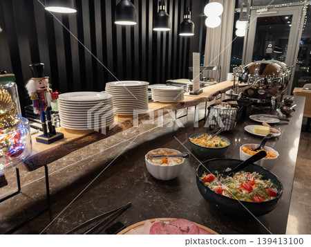 Buffet setup with fresh salad bowls and stacked white plates on a marble counter in a dining hall, reflecting a festive holiday hospitality atmosphere for hotel guests and luxury restaurant diners Buffet setup with fresh salad bowls and stacked white plates on a marble counter in a dining hall, reflecting a festive holiday hospitality atmosphere for hotel guests and luxury restaurant diners 139413100