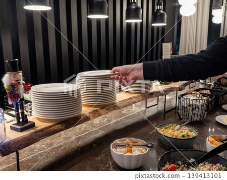 Person picking a plate from a stack at a hotel buffet, showcasing dining hospitality and catering services during holiday season with festive nutcracker decoration on a modern rustic wood counter top Person picking a plate from a stack at a hotel buffet, showcasing dining hospitality and catering services during holiday season with festive nutcracker decoration on a modern rustic wood counter top 139413101
