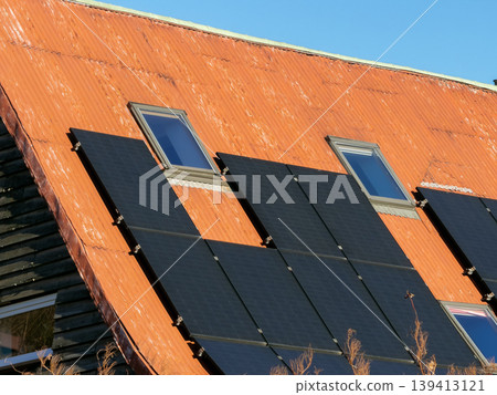 Black solar panels installed on a rusty red corrugated metal roof with attic windows, representing sustainable energy generation, home modernization, and eco-friendly power solutions for houses Black solar panels installed on a rusty red corrugated metal roof with attic windows, representing sustainable energy generation, home modernization, and eco-friendly power solutions for houses 139413121