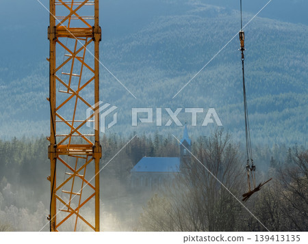 Tower crane mast and hanging construction materials framing a distant rural church in a misty forest landscape, representing the contrast between modern urban development and traditional architecture 139413135