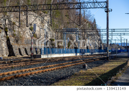 Railroad tracks run alongside a steep rocky cliff with overhead power lines and metal station fencing, capturing a scenic railway infrastructure journey through a mountainous forest landscape setting Railroad tracks run alongside a steep rocky cliff with overhead power lines and metal station fencing, capturing a scenic railway infrastructure journey through a mountainous forest landscape setting 139413149