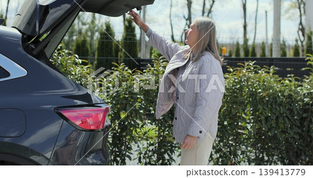 Senior woman puts bag in car trunk and closes it with button. Senior woman puts bag in car trunk and closes it with button. 139413779