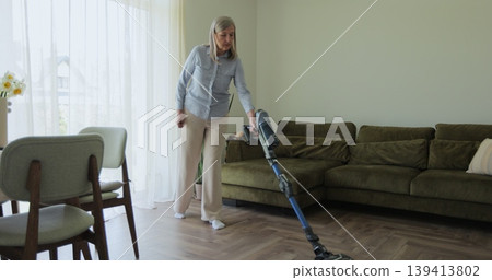 Older woman in casual clothes doing daily housekeeping chores , hand vacuuming carpet from dust using hoover. Older woman in casual clothes doing daily housekeeping chores , hand vacuuming carpet from dust using hoover. 139413802