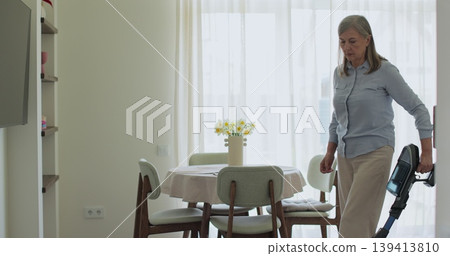 Senior woman cleans apartment by hand vacuuming carpet in living room. Concept of daily home routine. Senior woman cleans apartment by hand vacuuming carpet in living room. Concept of daily home routine. 139413810