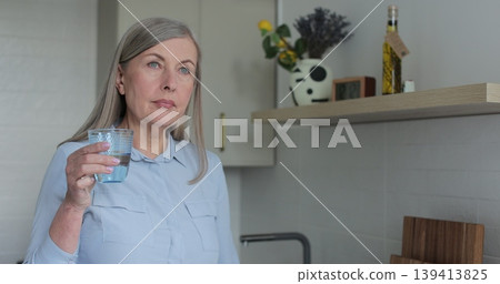 Senior mature woman drinking a glass of water in kitchen at home. Elderly, lifestyle, hydration, wellness, relaxation, home. 139413825