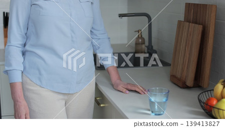 Senior mature woman drinking a glass of water in kitchen at home. Elderly, lifestyle, hydration, wellness, relaxation, home. Senior mature woman drinking a glass of water in kitchen at home. Elderly, lifestyle, hydration, wellness, relaxation, home. 139413827