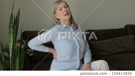 Mature woman feeling ache lower back pain on sofa at home. Upset elderly retired woman massages painful lower back muscles. Senior lady feel spasm. Mature woman feeling ache lower back pain on sofa at home. Upset elderly retired woman massages painful lower back muscles. Senior lady feel spasm. 139413850