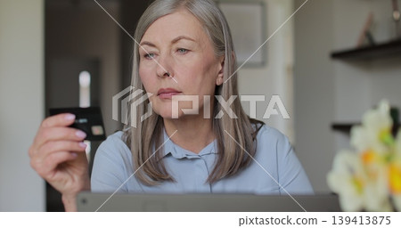 Old woman using laptop at home in the living room. Elderly woman doing shopping online and pay bill by credit bank card using her laptop. Online purchase. Old woman using laptop at home in the living room. Elderly woman doing shopping online and pay bill by credit bank card using her laptop. Online purchase. 139413875