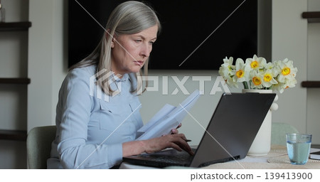 Senior woman sits at table viewing papers and utility bills. Senior woman sit with laptop and paper document. Senior woman sits at table viewing papers and utility bills. Senior woman sit with laptop and paper document. 139413900