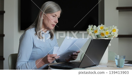 Senior woman sits at table viewing papers and utility bills. Senior woman sit with laptop and paper document. Senior woman sits at table viewing papers and utility bills. Senior woman sit with laptop and paper document. 139413903
