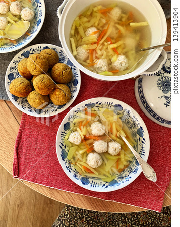 A bowl of vegetable soup with chicken meatballs is served alongside chickpea buns on a table. Fresh ingredients create a hearty meal for breakfast or lunch A bowl of vegetable soup with chicken meatballs is served alongside chickpea buns on a table. Fresh ingredients create a hearty meal for breakfast or lunch 139413904