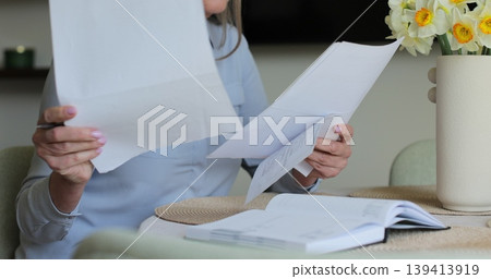 Unrecognisable elderly lady looking through utility bills putting them on round coffee table and typing on laptop. Unrecognisable elderly lady looking through utility bills putting them on round coffee table and typing on laptop. 139413919