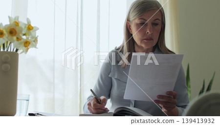 Close up older gray-haired woman sits at a table in the living room, calculating bills and taxes and taking notes. 139413932